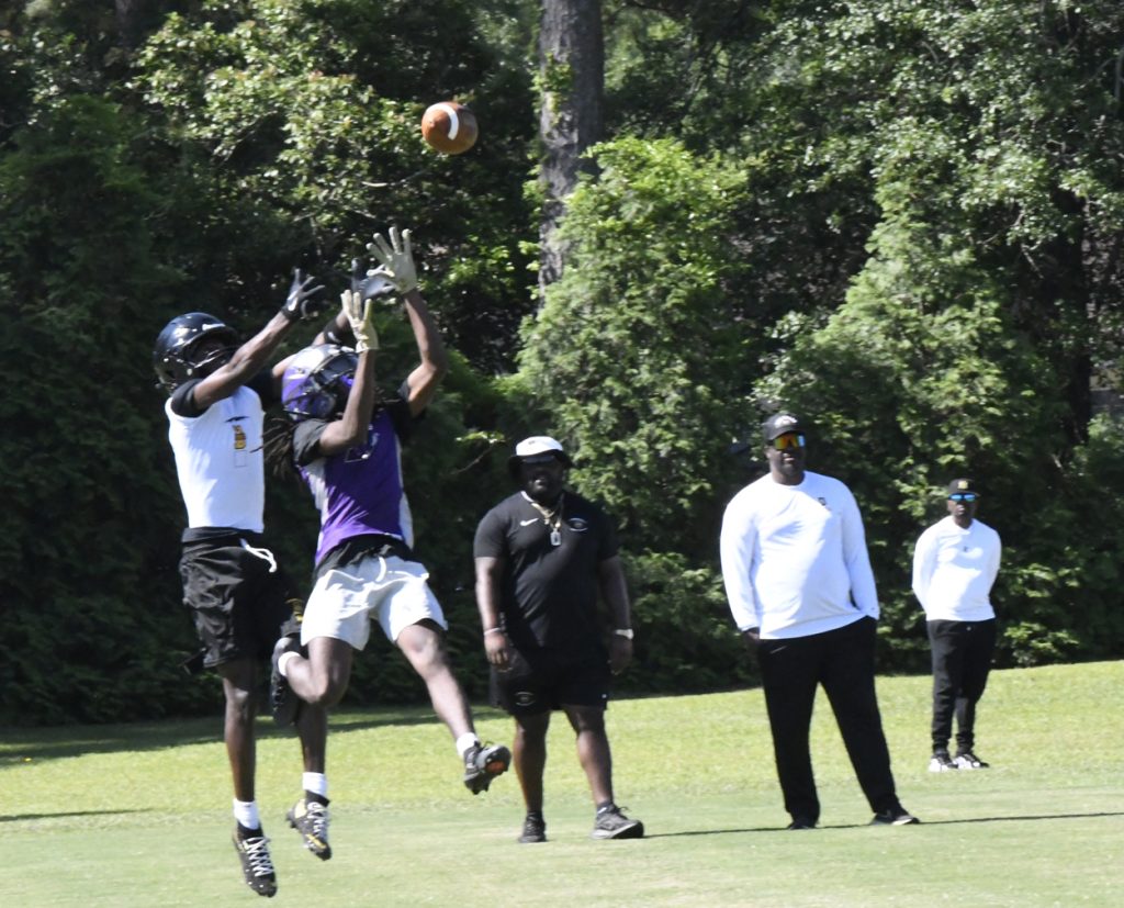 Two football players jump to catch a ball during practice, while coaches watch from the sidelines on a grassy field surrounded by trees.