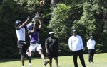 Two football players jump to catch a ball during practice, while coaches watch from the sidelines on a grassy field surrounded by trees.