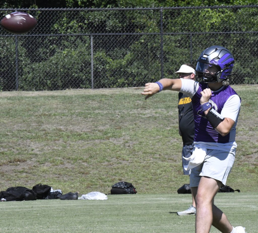 Football player in purple helmet and vest throws a football during practice on a grassy field, with coach and equipment visible in the background.