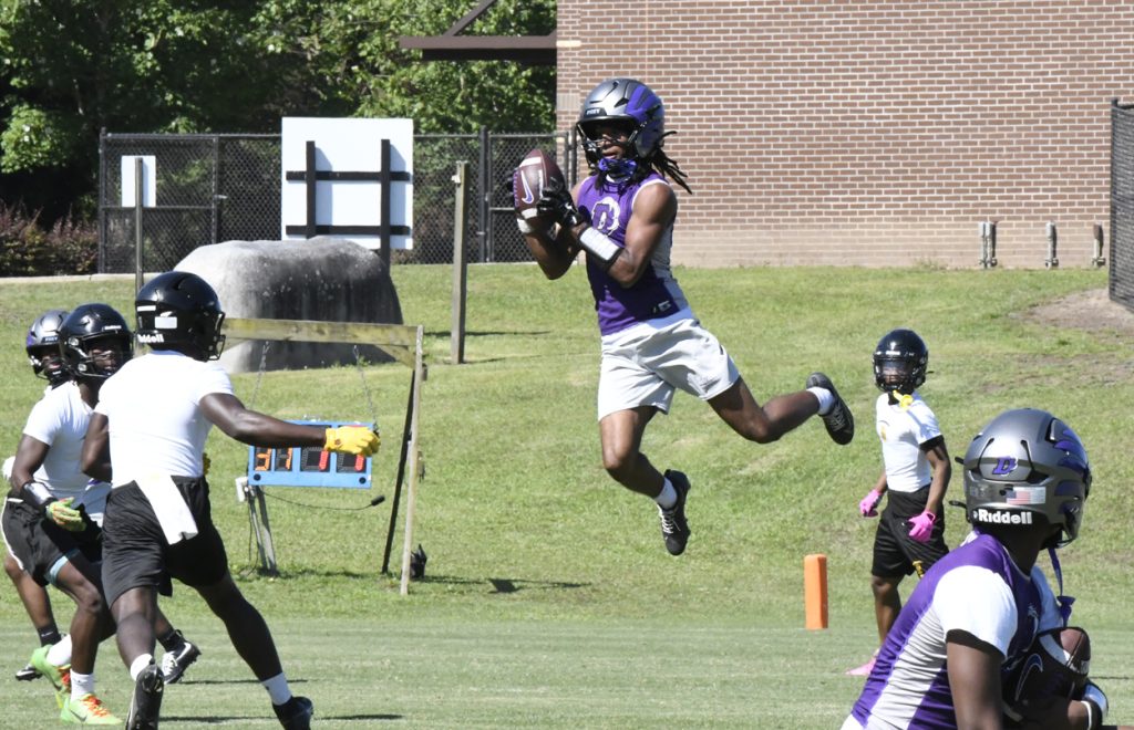 A football player in a purple jersey jumps to catch a pass during a game, with defenders nearby on a grassy field.