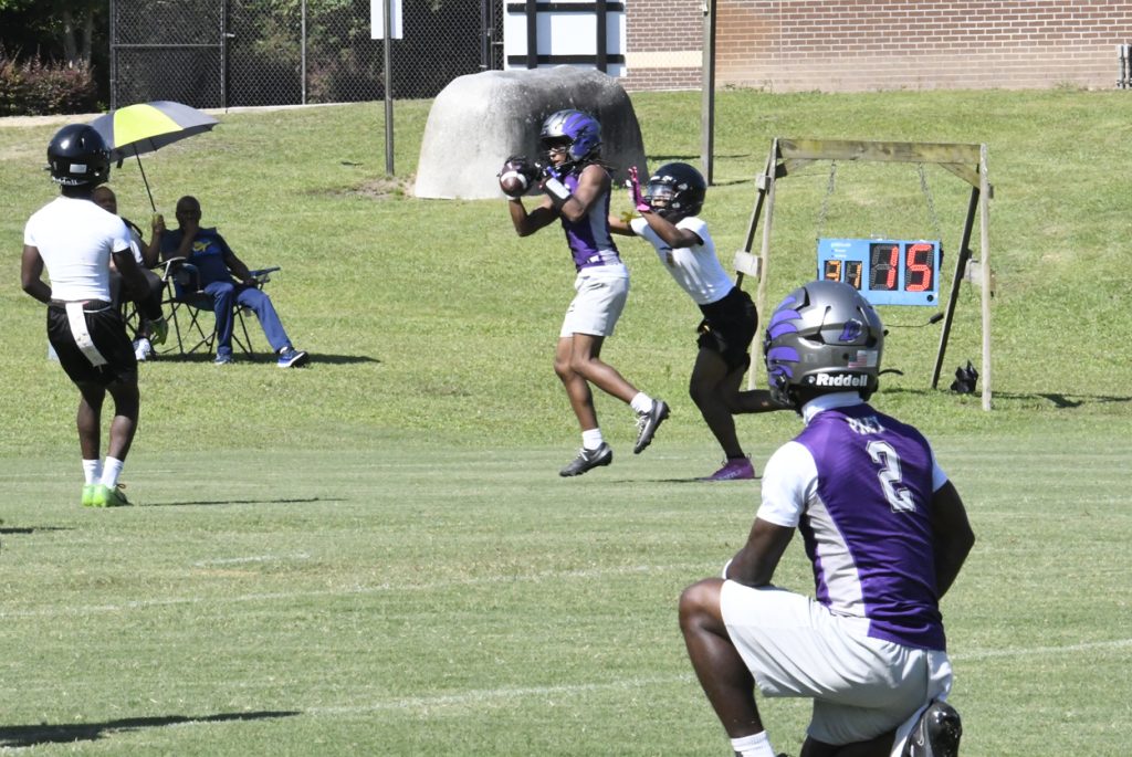 A football player in purple catches a pass while being closely defended, with a teammate and spectators visible on a grassy field.