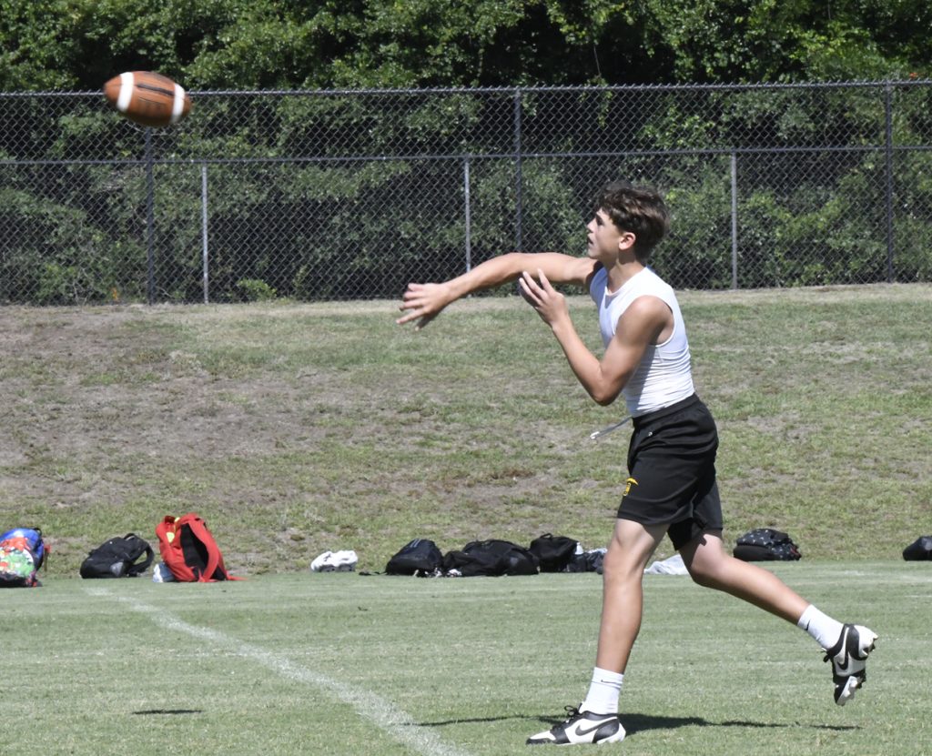 A young athlete in a sleeveless shirt throws a football on a grassy field, with bags and a fence visible in the background.