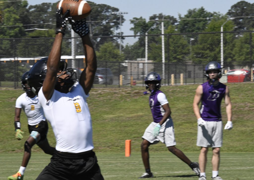 A football player in a white shirt catches a pass during a game, while other players in purple jerseys watch on a sunny outdoor field.