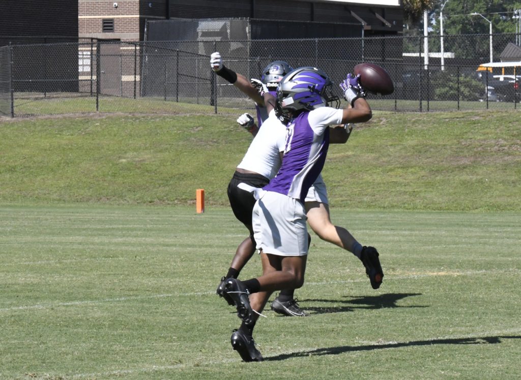 Two football players compete for a catch on a grassy field, both reaching for the ball, with a building and fence in the background.