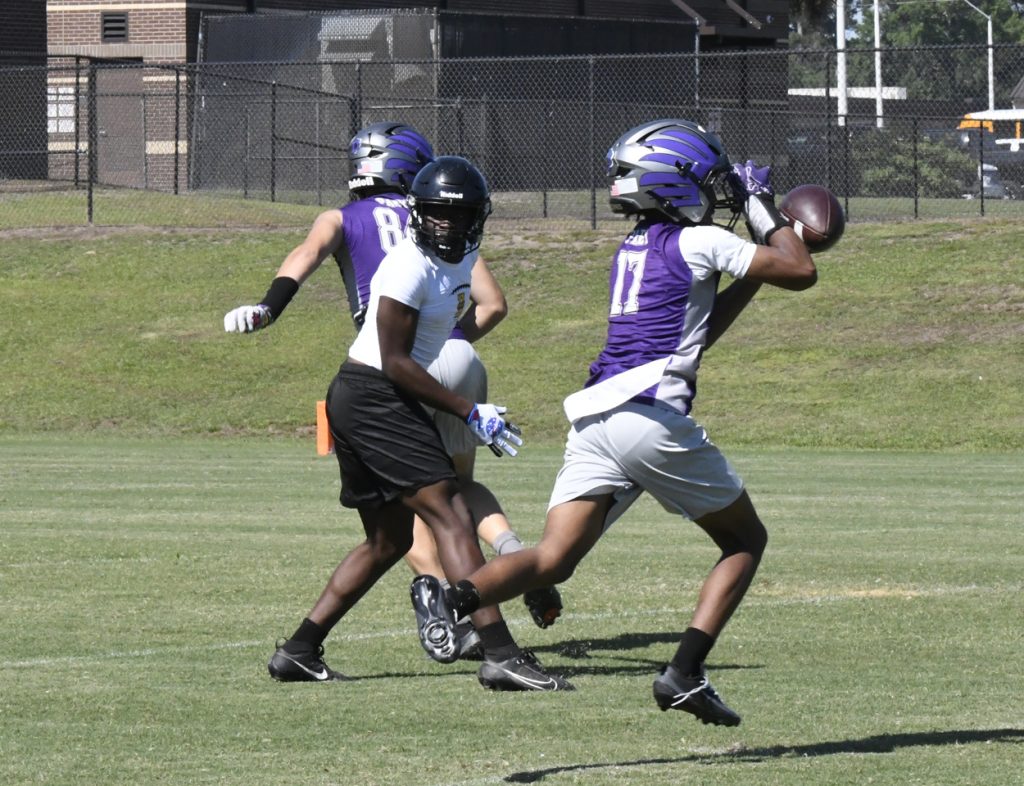 Football players practice on a grassy field, one player wearing a purple jersey catches the ball while others defend.