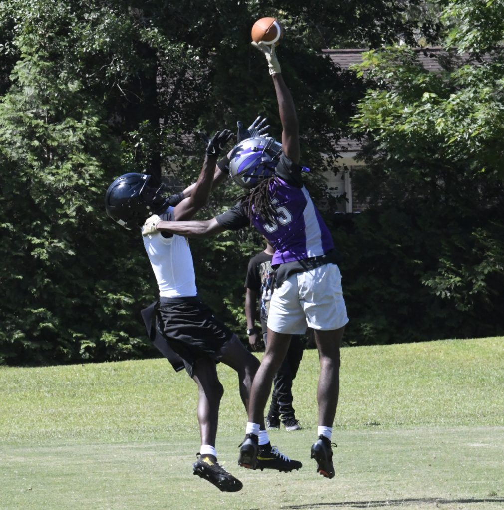 Two football players jump to catch a ball during a game on a grassy field, both wearing helmets and athletic gear, with trees visible in the background.