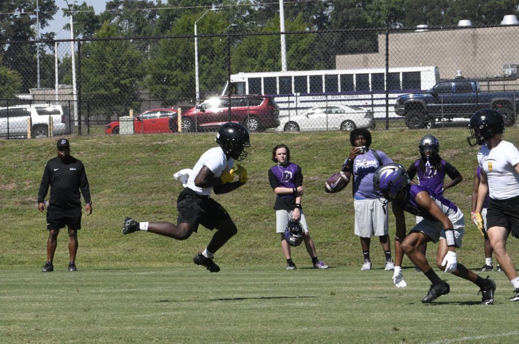 A football player jumps to catch a ball during a practice session on a grassy field, with coaches and players watching in the background.