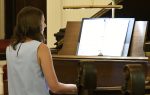 A person with long hair plays piano, reading sheet music on a stand in a softly lit room.