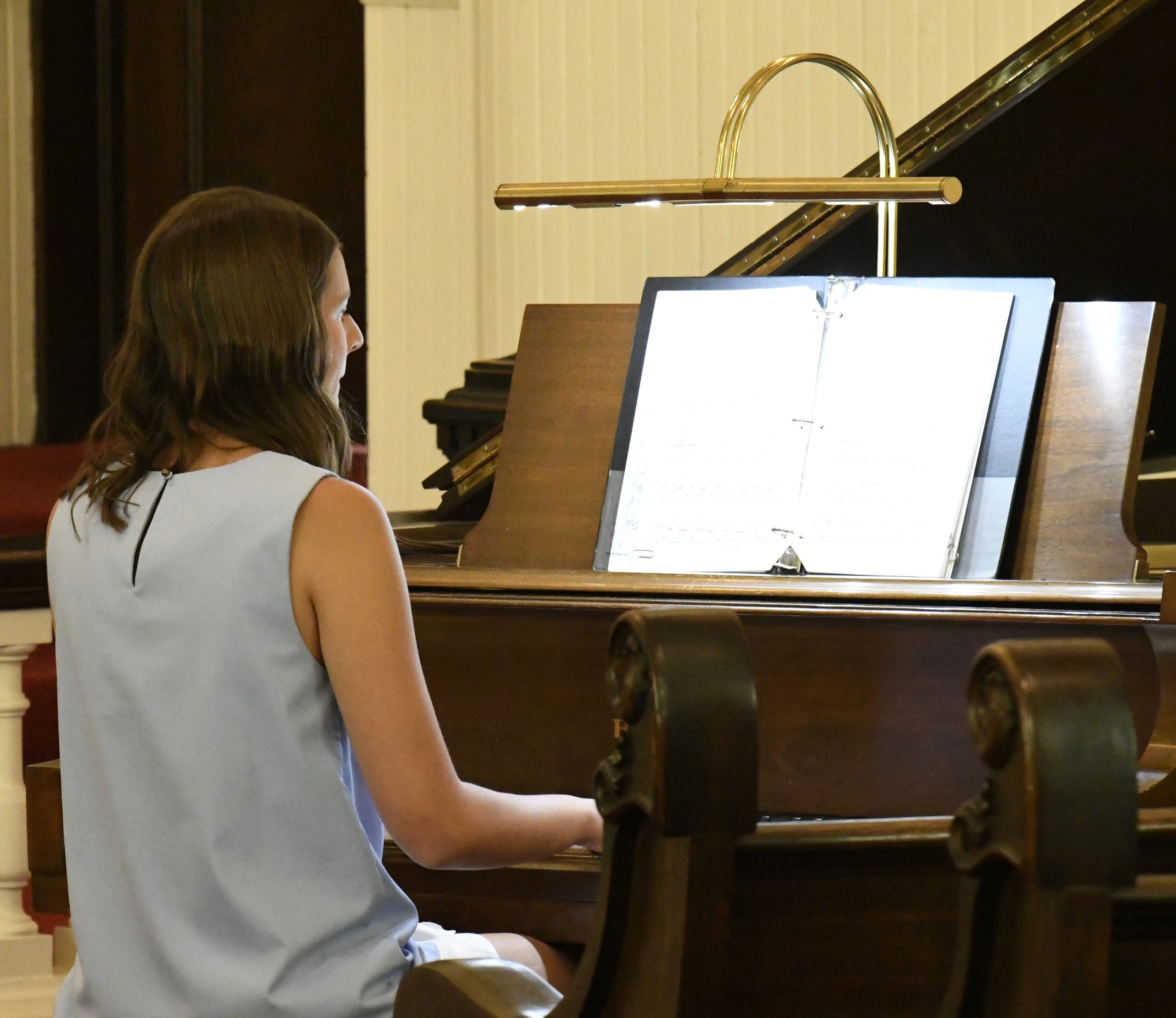 A person with long hair plays piano, reading sheet music on a stand in a softly lit room.