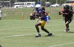 Football player in blue jersey and yellow shorts preparing to throw a football during outdoor practice on a grassy field.