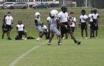 Football players in helmets and gear practice on a grassy field, with others watching in the background near a parking lot and school buses.