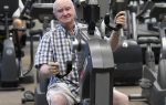 elderly man in a plaid shirt exercises on an arm cycling machine at a gym, surrounded by other fitness equipment.