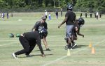Football players in helmets and athletic gear practice drills on a grassy field with cones, while a coach observes and instructs them during a training session.