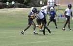 Football players in helmets and pads engage in a tackle during a practice or scrimmage on a grassy field.
