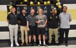Six men stand together indoors; the central blonde man holds a trophy. Black-and-gold balloons and a logo backdrop appear behind them, suggesting an award ceremony or team celebration.