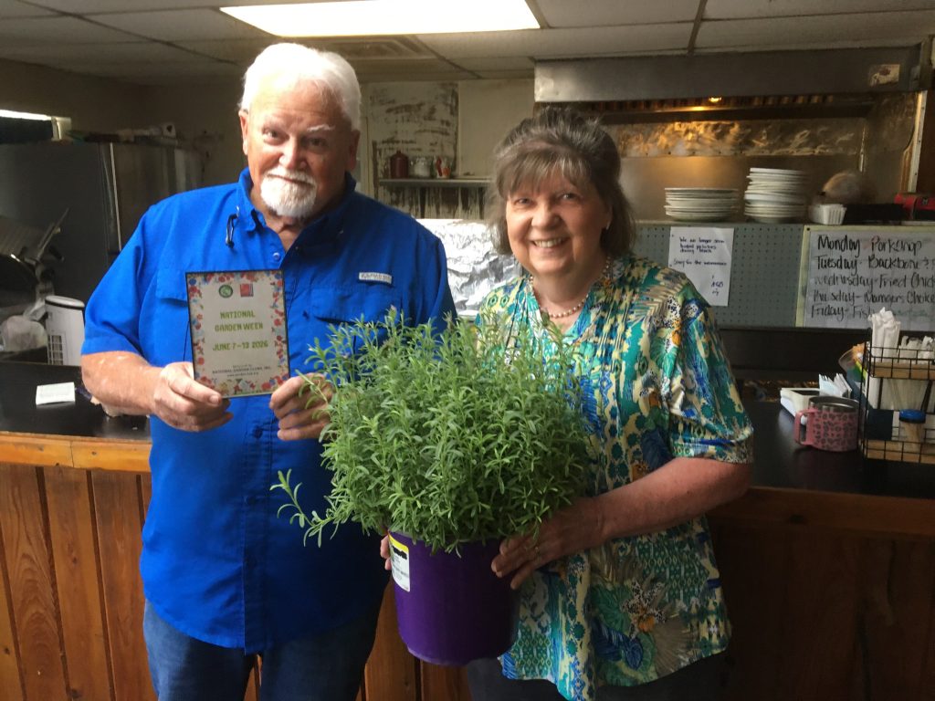 older man holds a National Garden Week sign while a smiling woman holds a potted plant inside a kitchen or restaurant setting.