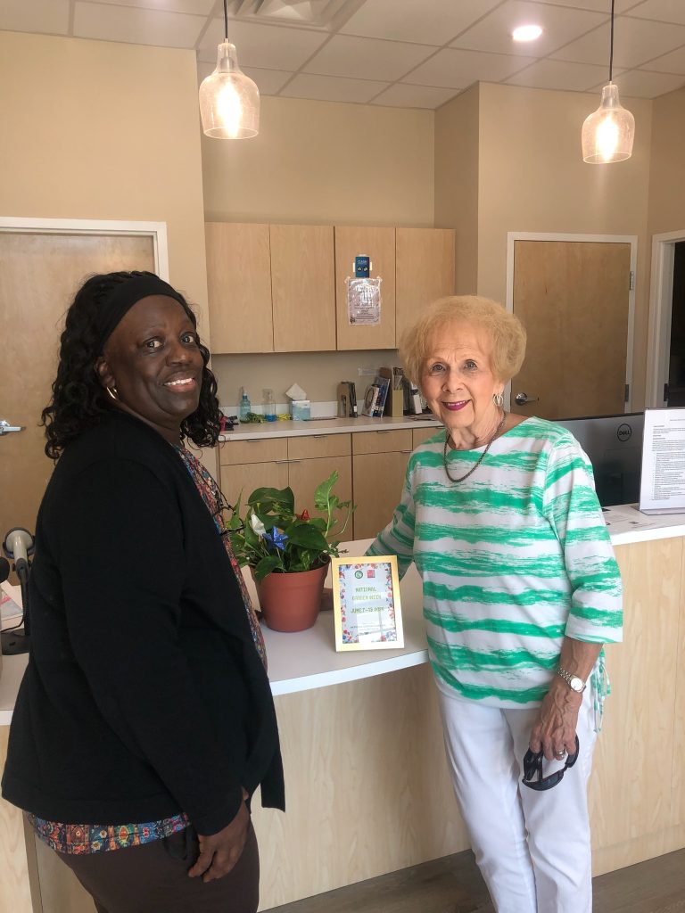 Two women standing and smiling in an office lobby by a counter with a potted plant and a framed sign about "National Garden Week."