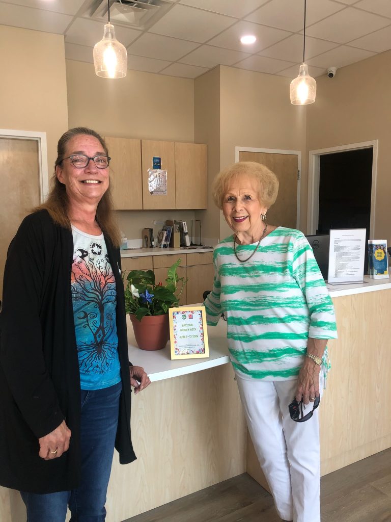 Two women smiling in a brightly lit office lobby, standing beside a potted plant and a framed sign on the counter.