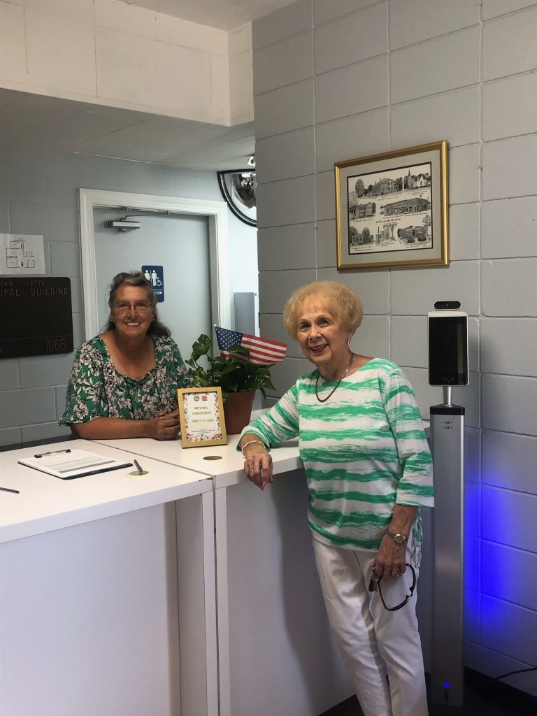 Two elderly women smiling in an office lobby, one standing, one sitting at a desk with papers, plant, framed artwork, and an American flag visible.