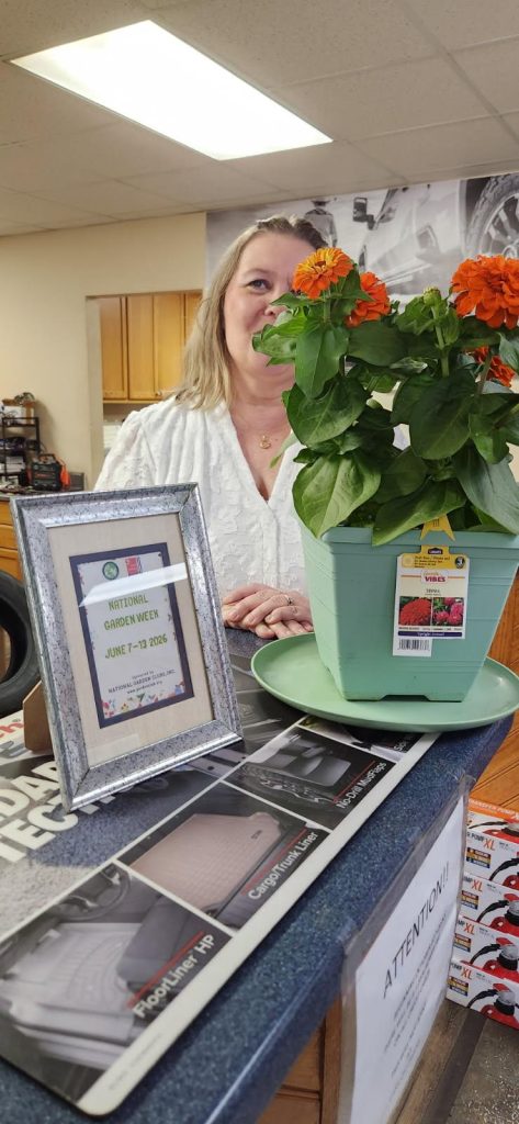 A woman stands behind a counter with a potted orange zinnia plant and a framed National Garden Week sign dated June 7-13, 2026.