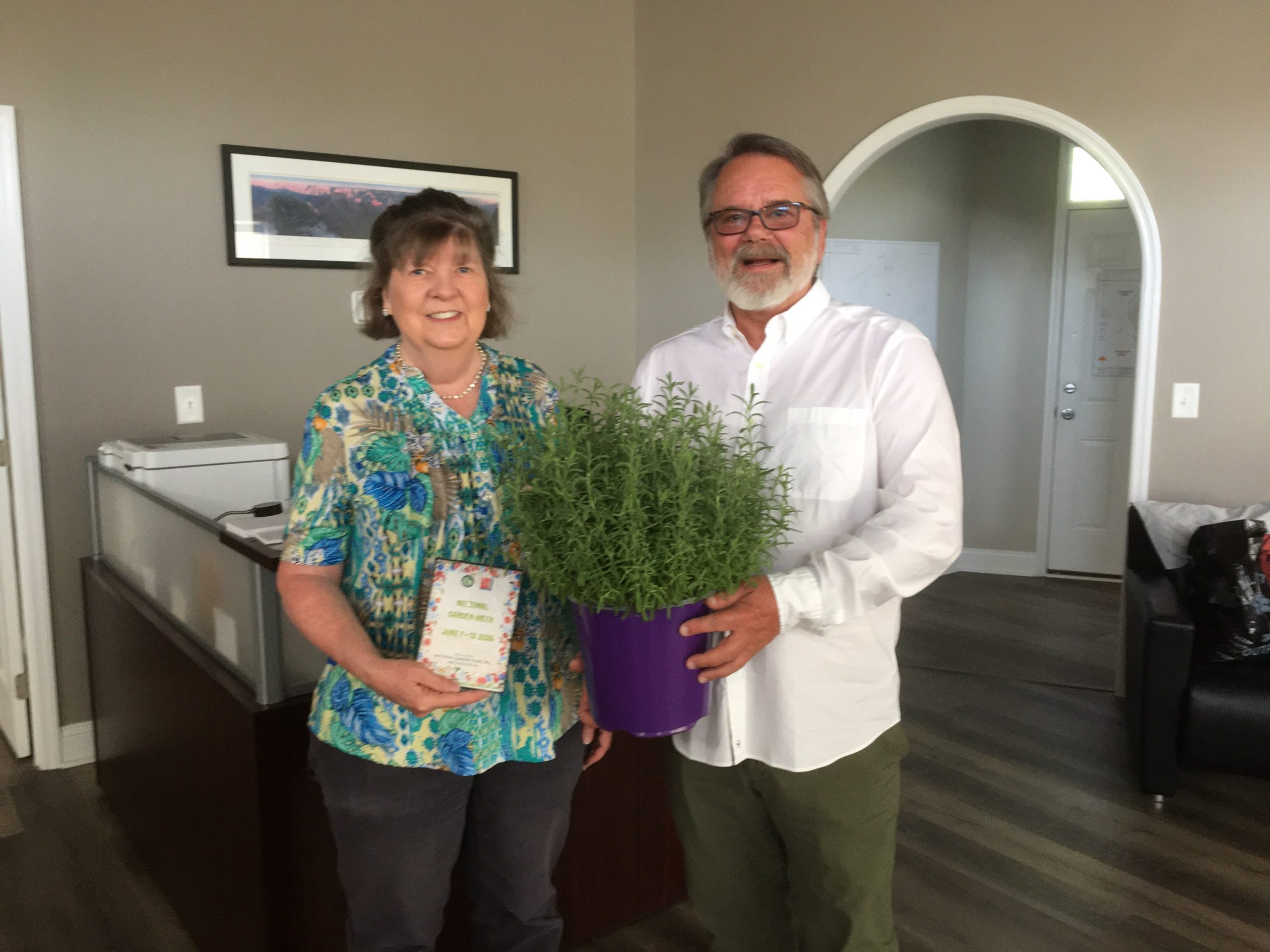 A woman and man stand indoors, smiling, holding a potted plant in a purple container and a greeting card.