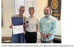 Three people standing indoors, two women holding an award certificate and a box, a man stands beside them, all smiling, with a Rotary Club banner in the background.