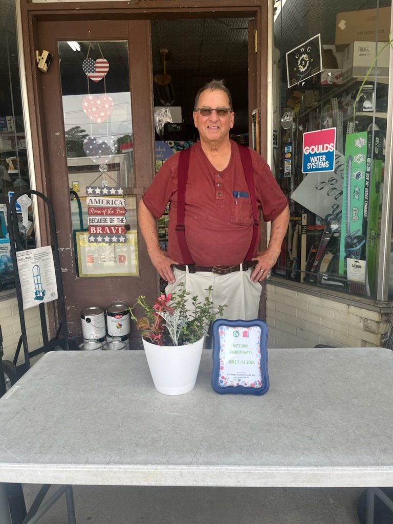 Man in red shirt stands behind a table with a potted plant and a "National Garden Week" sign in front of a store entrance.