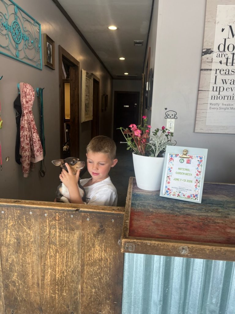A young boy holds a small dog behind a wooden counter, with a potted plant and a "National Garden Week" sign visible nearby.