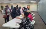 People sitting at a long table indoors, handing out materials to others who are lined up and waiting in a spacious, well-lit room.