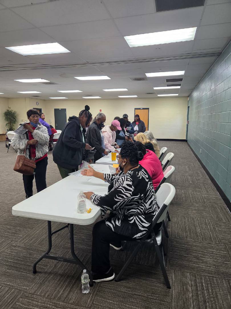 People sitting at a long table indoors, handing out materials to others who are lined up and waiting in a spacious, well-lit room.