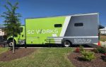 A bright green and gray SC Works mobile unit parked on grass under a clear blue sky, with landscaping in the foreground.