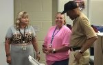 Three adults standing indoors, two women and one man, talking and smiling. One woman wears a cheetah print top, another a pink shirt, the man wears a tan shirt and black cap.