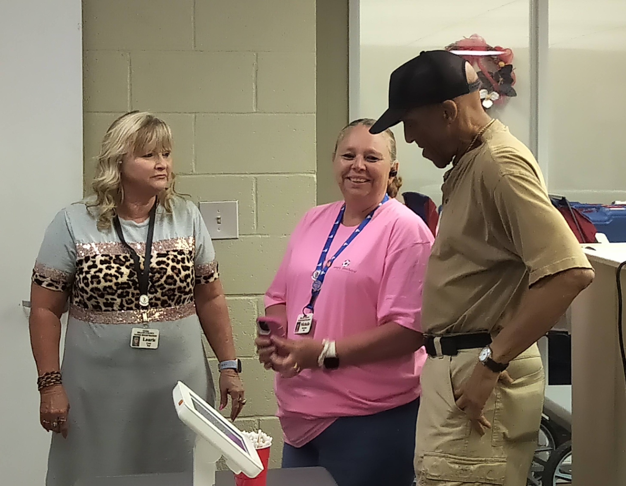 Three adults standing indoors, two women and one man, talking and smiling. One woman wears a cheetah print top, another a pink shirt, the man wears a tan shirt and black cap.