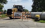 Large construction vehicle behind road closed sign, orange and white barricades, and concrete pipes blocking a street under repair, with trees and blue sky in the background.
