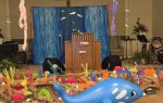A church stage decorated with an underwater theme, featuring colorful coral, paper fish, blue backdrop, and a large blue inflatable whale in front of the podium.