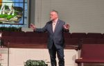 A man in a suit gestures while speaking in a church with a stained glass window and empty pews in the background.