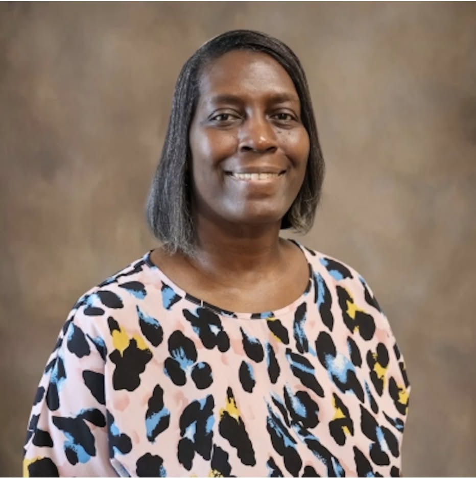 Smiling woman with straight shoulder-length hair, wearing a colorful animal print blouse, posed in front of a plain brown background.