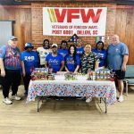 Group of people standing behind a table with donated food items at a VFW post, decorated with patriotic colors and a VFW banner in the background.
