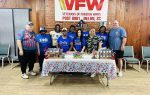 Group of people standing behind a table with donated food items at a VFW post, decorated with patriotic colors and a VFW banner in the background.