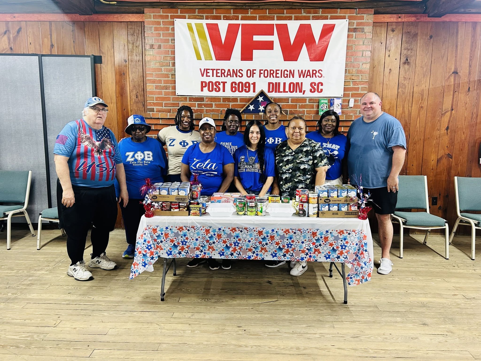 Group of people standing behind a table with donated food items at a VFW post, decorated with patriotic colors and a VFW banner in the background.