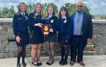 Five people in formal attire, four women in blue FFA jackets and one man in a suit, posing outside with an award plaque.