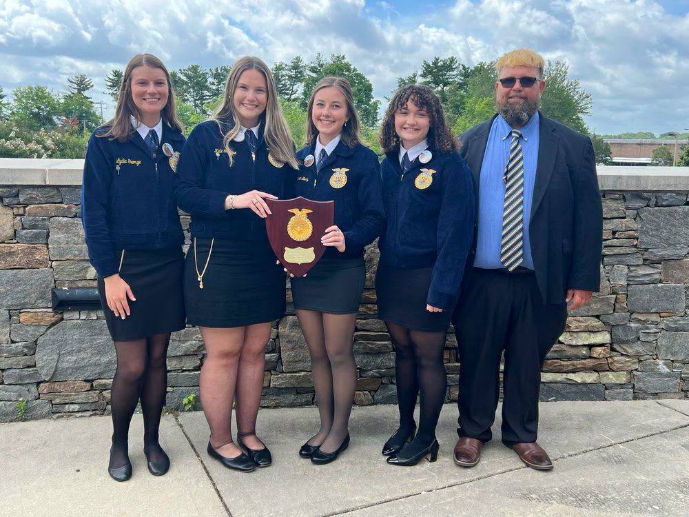 Five people in formal attire, four women in blue FFA jackets and one man in a suit, posing outside with an award plaque.