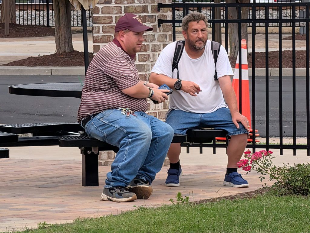 Two men sitting on an outdoor bench near a brick wall, one wearing a striped shirt and cap, the other in a white t-shirt and shorts.