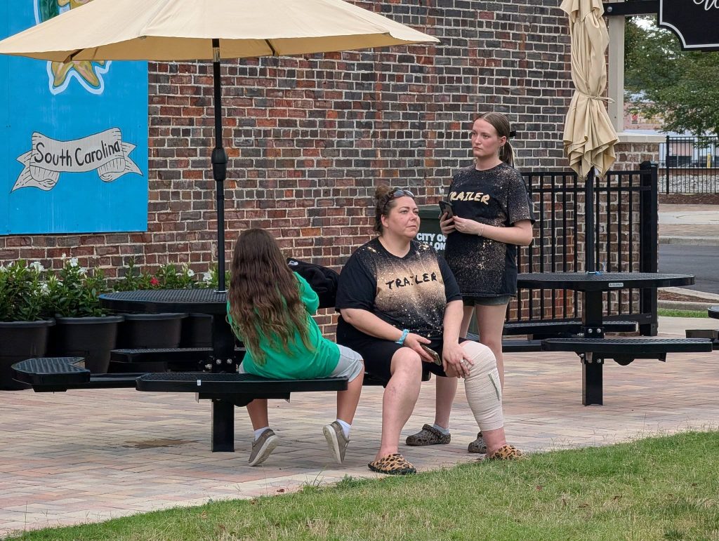 Three people relaxing outside on black metal picnic tables, with a brick wall and "South Carolina" sign in the background. One person has a bandaged leg.