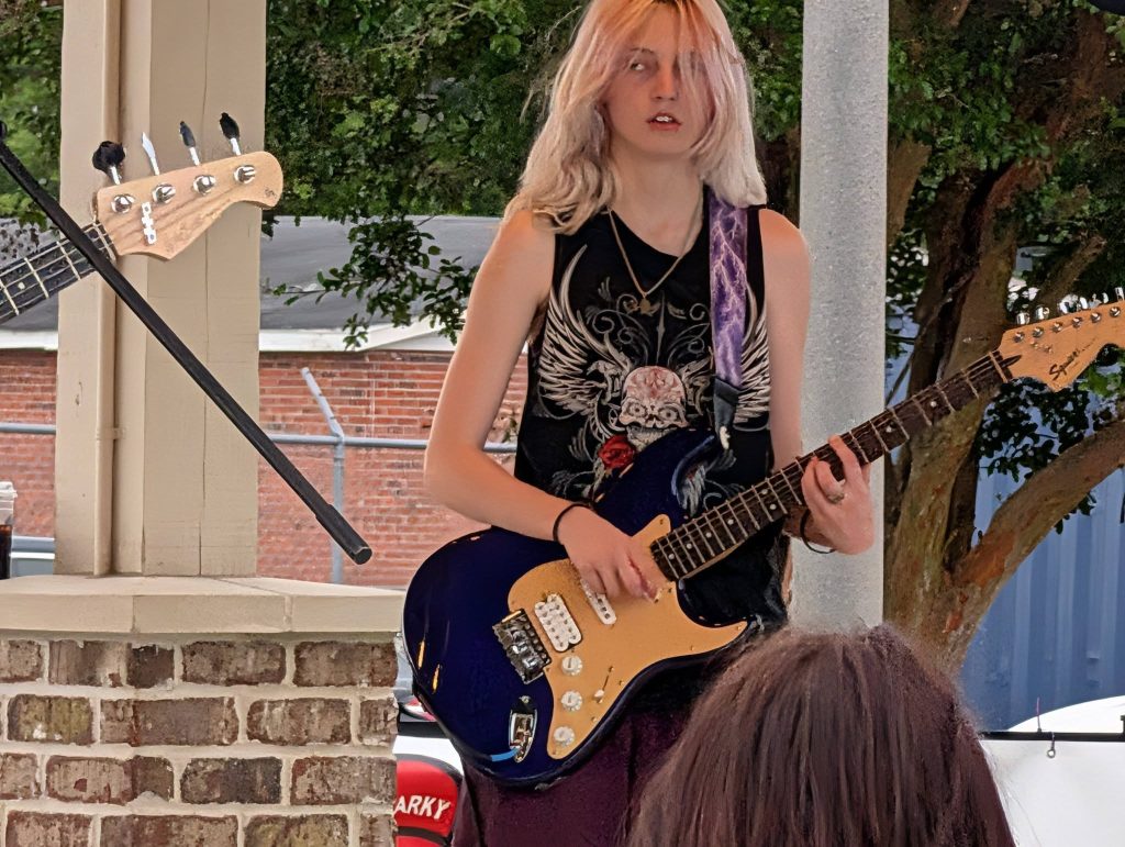 Person with long blonde hair playing an electric guitar on an outdoor stage, wearing a black shirt with a skull design, trees and brick wall in background.