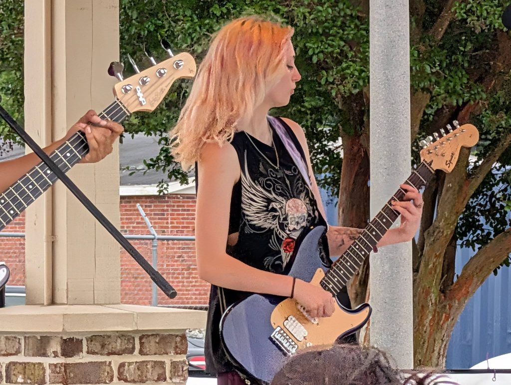 Young woman with dyed hair playing an electric guitar outdoors, wearing a black shirt with a skull and wings design. Another musician with a bass guitar is partially visible.