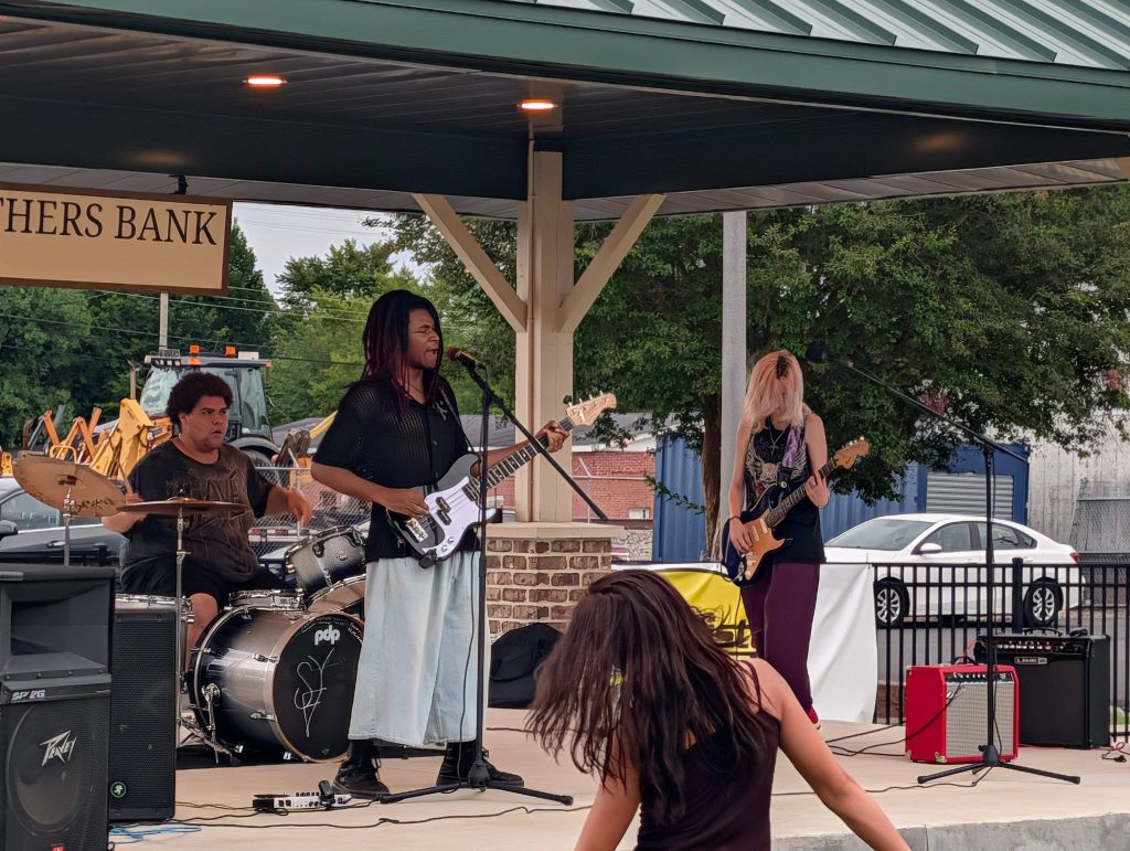 A three-piece band performs on an outdoor stage with drums, bass, and guitar while a person dances in the foreground.