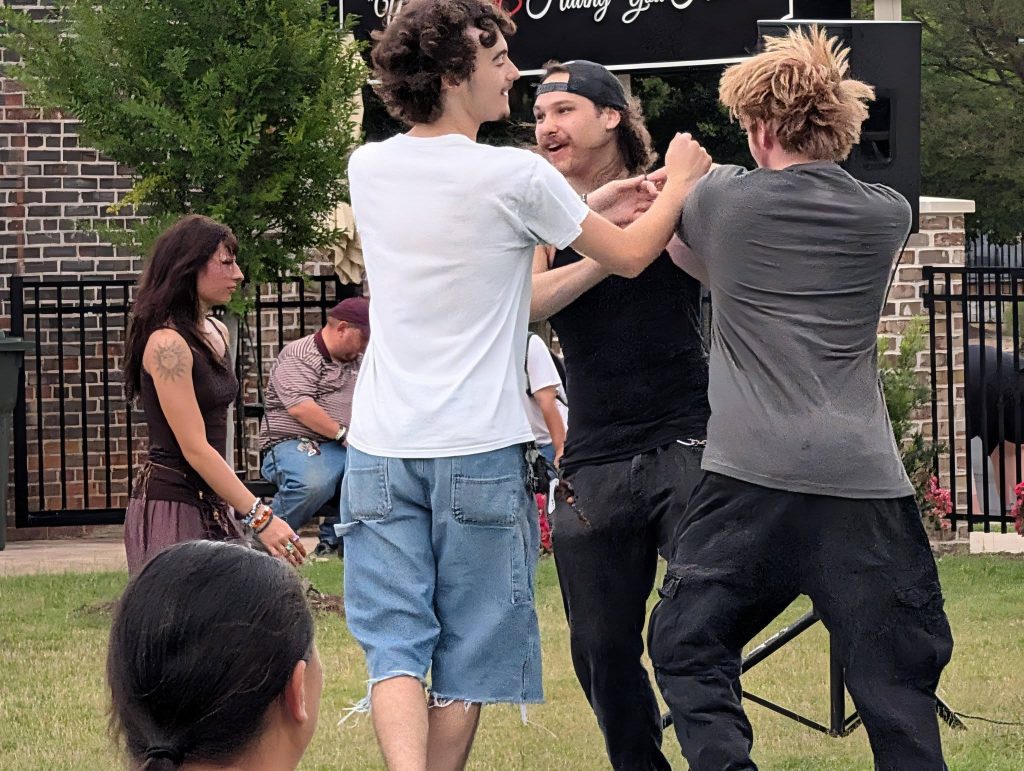 Three young men interact animatedly while a woman with a tattoo watches nearby; another person sits on a bench in the background.