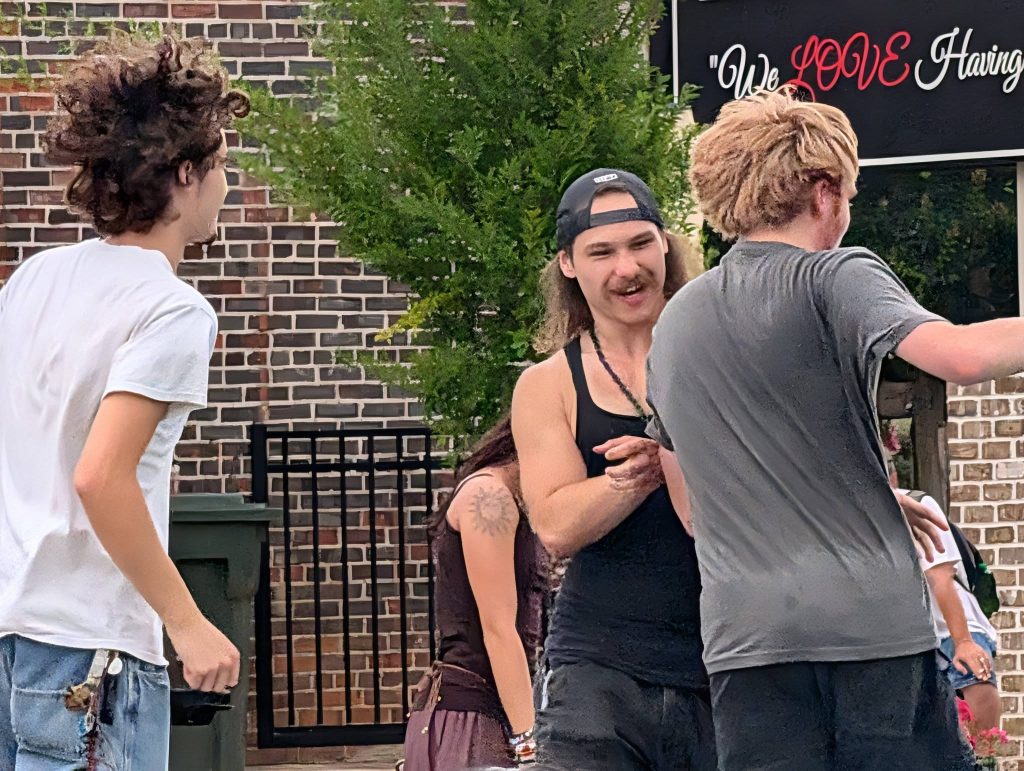 Three young people interact outdoors near a brick wall, with one wearing a backward cap and another with curly hair. A sign in the background reads “We LOVE Having.”