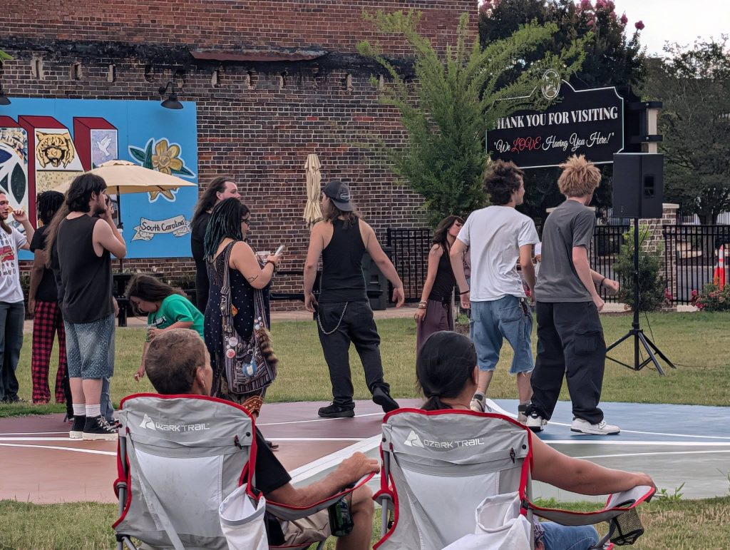 People gather and socialize outdoors near a colorful mural and a “Thank You For Visiting” sign, with two seated individuals in the foreground on lawn chairs.